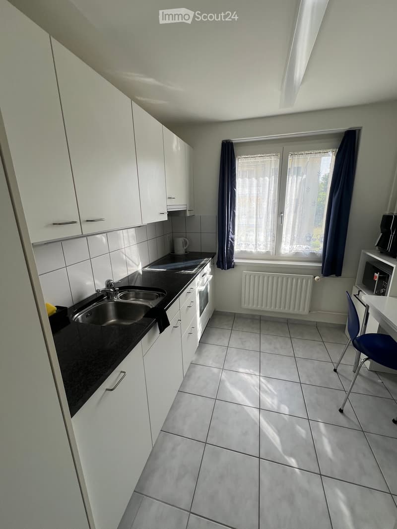 Modern kitchen with white cabinets, black countertop, stainless steel sink, and a small table with a blue chair.