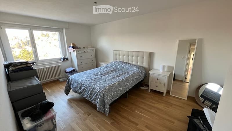 Bright bedroom with wooden flooring, large window, upholstered headboard bed, nightstand, dresser, and mirror. Storage boxes and folded clothes are visible.