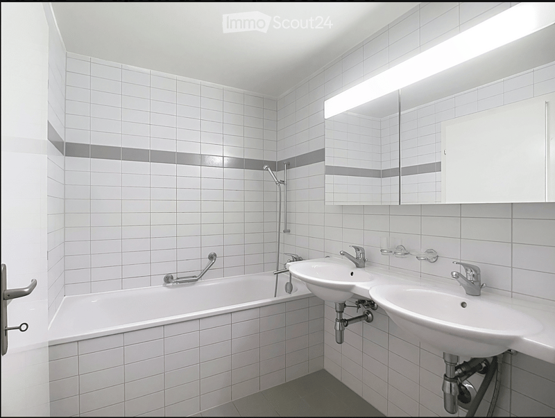 Modern bathroom with white tiled walls and floor, featuring a bathtub, two sinks with chrome faucets, a large mirror with built-in lighting, and a white cabinet.