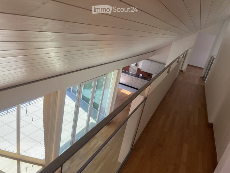 Spacious hallway with wooden flooring, modern railing, and large windows overlooking a tiled outdoor area. The ceiling is slanted with wooden paneling, and a small kitchen area is visible in the background.