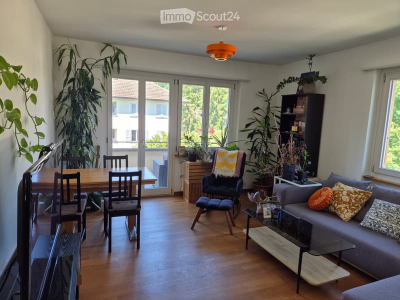 Bright living room with wooden flooring, large windows, and sliding glass doors leading to a balcony. Features a grey sofa with patterned cushions, a glass coffee table, a wooden dining table with chairs, a blue armchair with a blanket, and a bookshelf wi