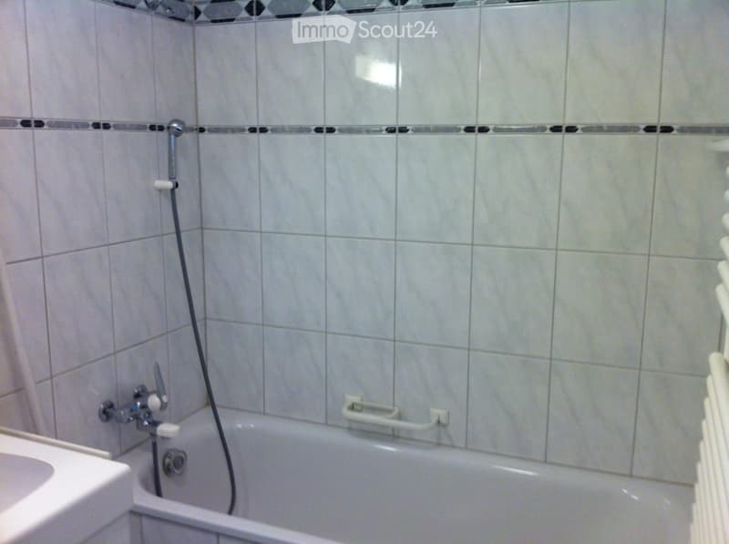 Bathroom with a bathtub, a shower hose, a faucet, and a handrail. The walls are tiled with light-colored tiles and a decorative border.