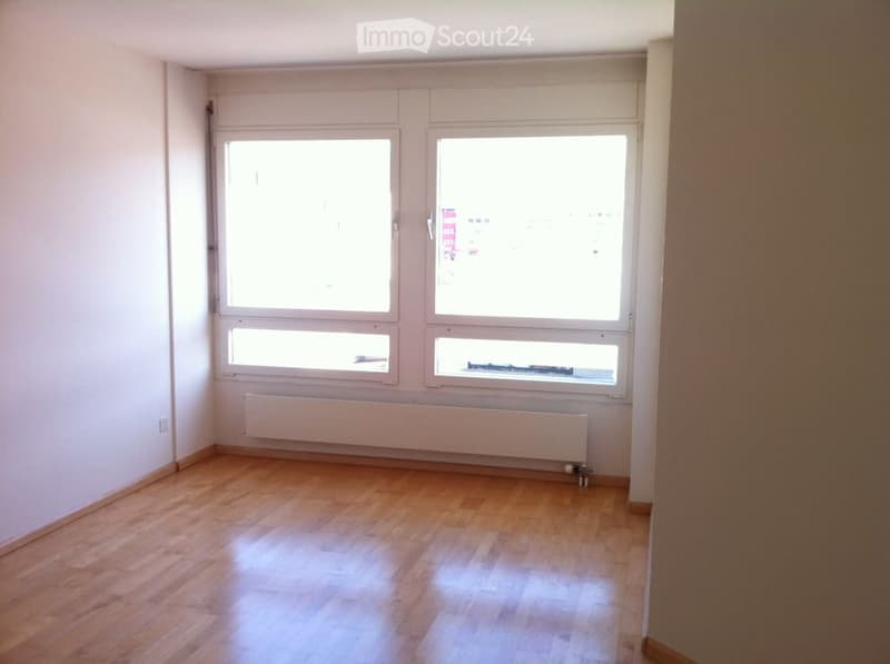 Empty room with wooden flooring, white walls, and large windows allowing natural light. A radiator is visible below the windows.