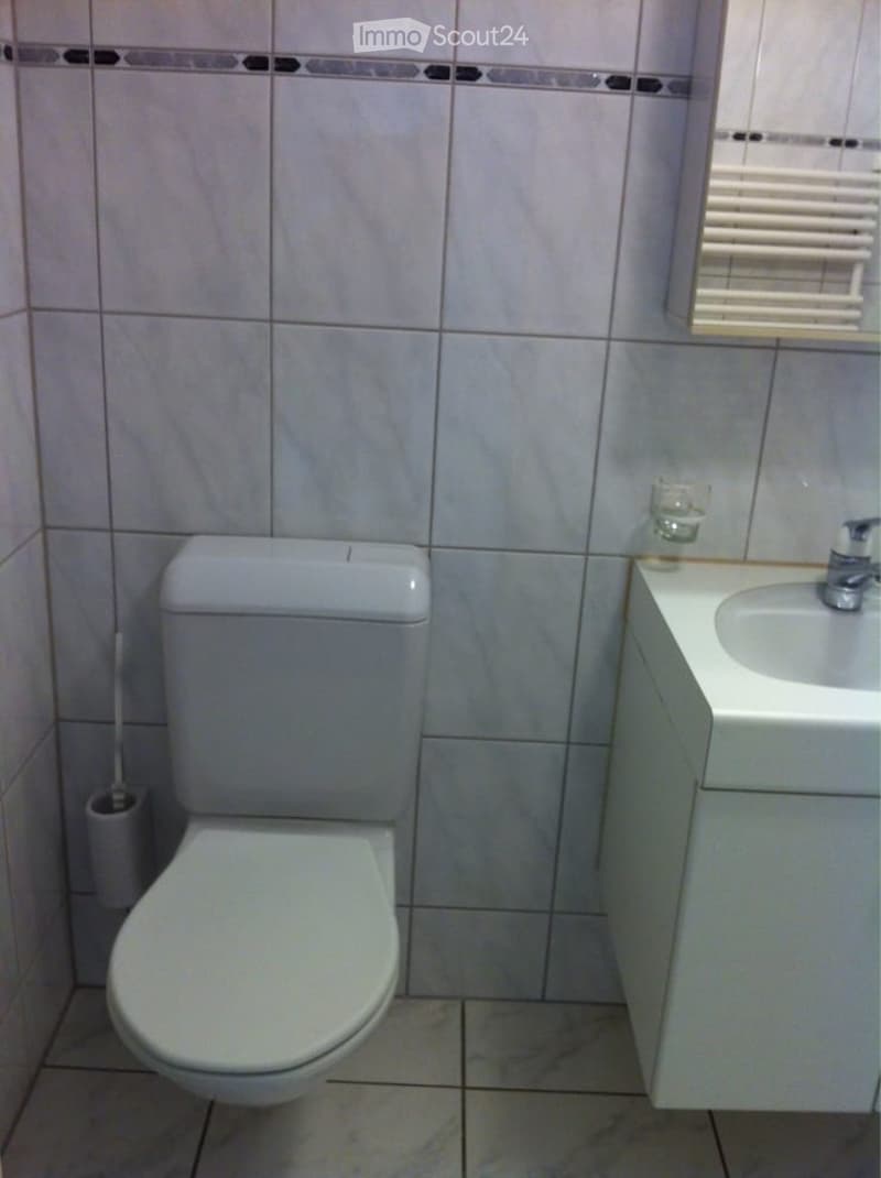 A small bathroom with a toilet, sink, and mirror. The walls are tiled with a decorative border, and a cup is placed on the sink counter.