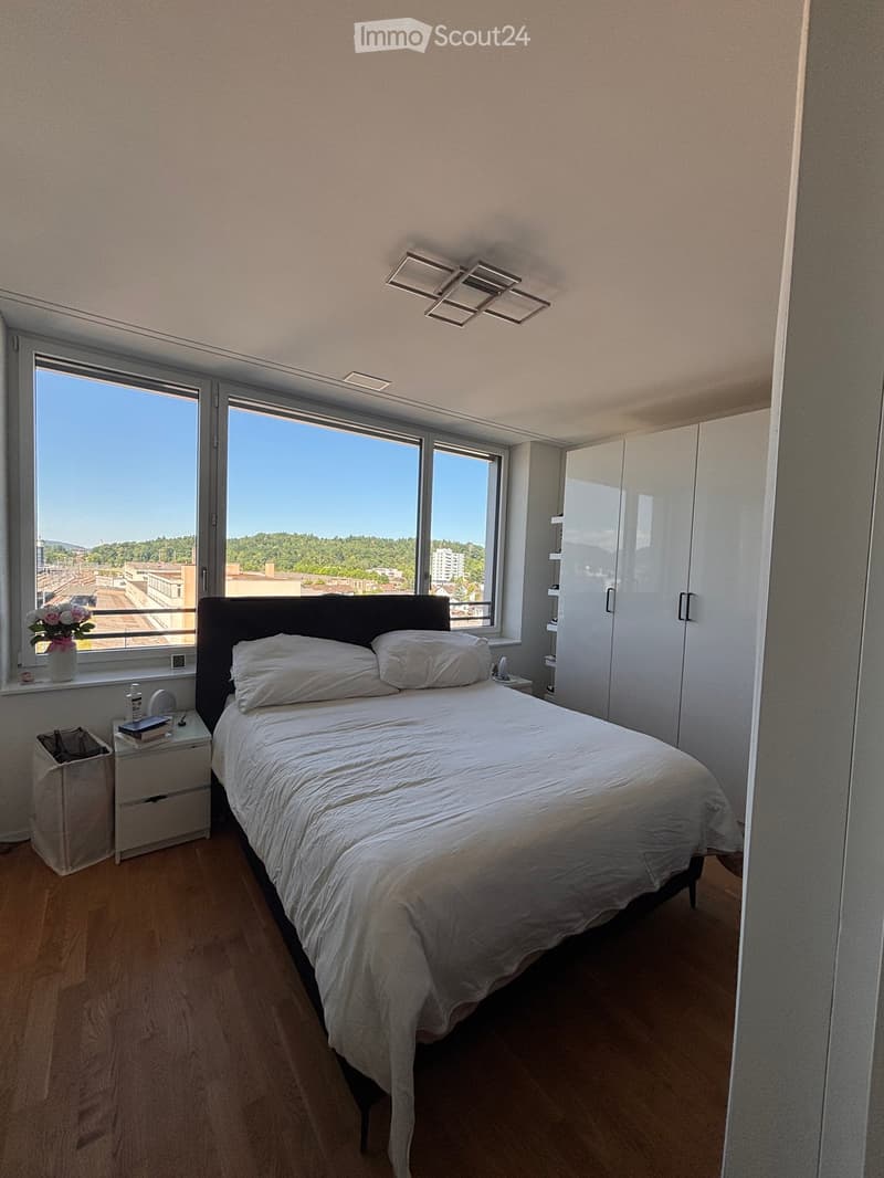 Modern bedroom with a large bed, nightstand, books, vase with flowers, and a wardrobe. Large windows offer a scenic view.