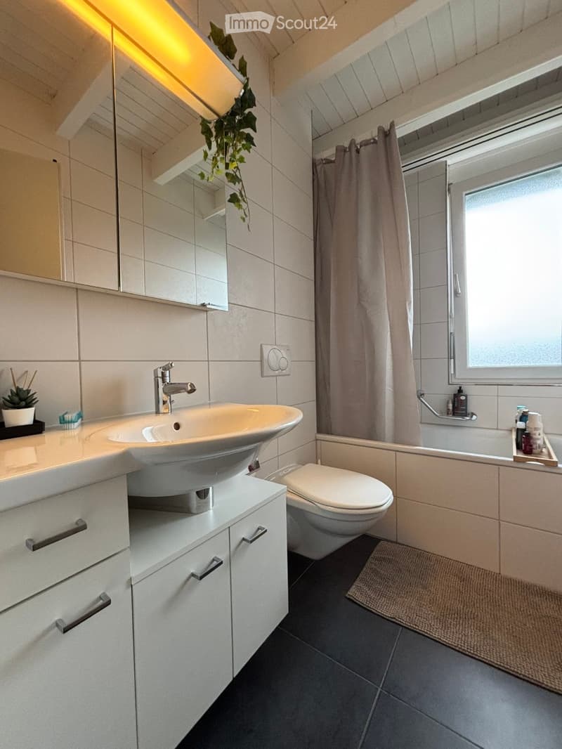 Modern bathroom with white tiles, a large sink with a modern faucet, a toilet, a bathtub with a shower curtain, and a window providing natural light. The space includes a cabinet with drawers, a mirror, and a small plant on the counter.