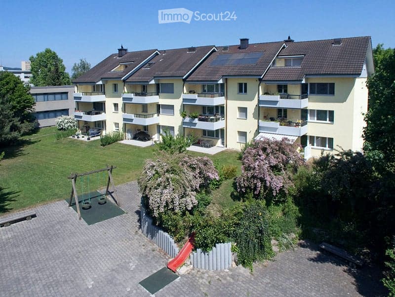 Multi-story residential building with balconies, solar panels on the roof, and a landscaped garden area with a swing set and slide.