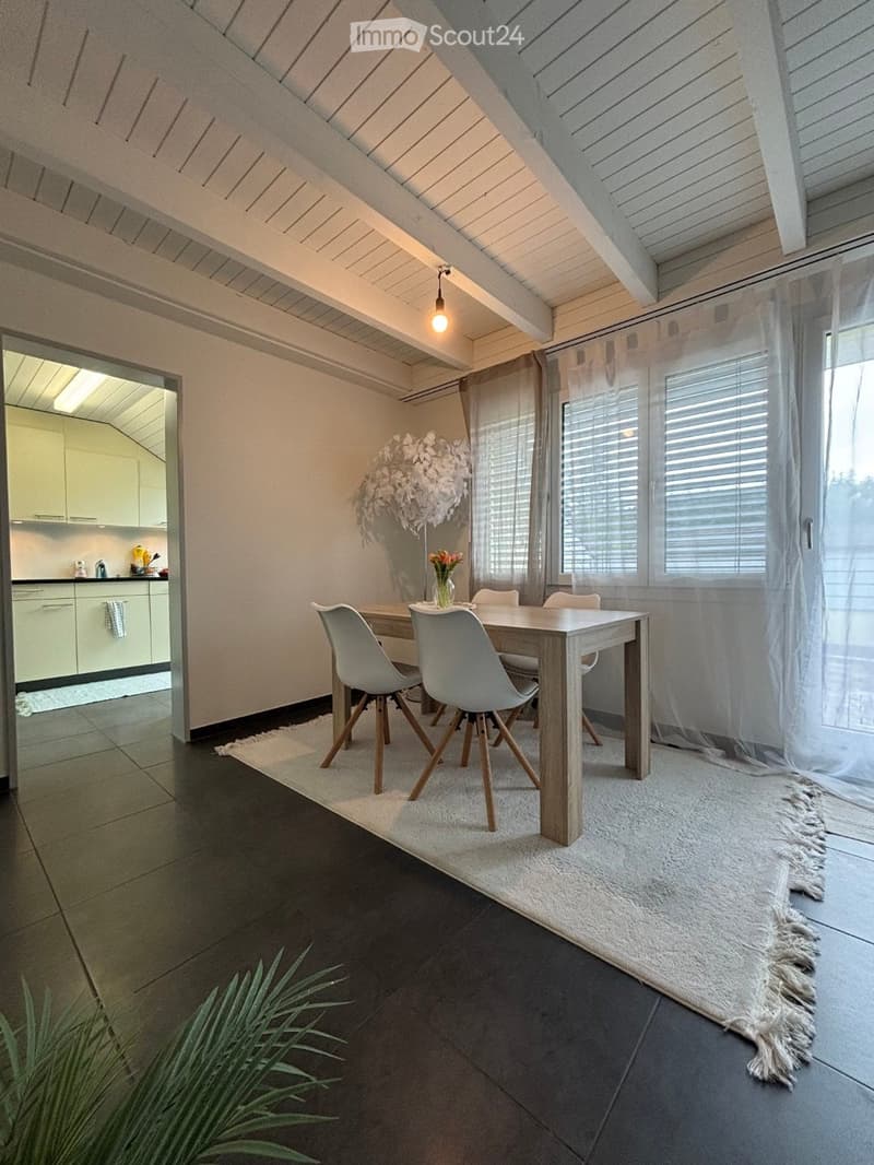Modern dining area with a wooden dining table, four grey chairs, a white rug, and a kitchen area visible in the background.