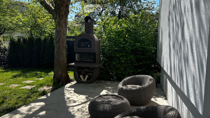 Outdoor patio area with a wood-fired oven, several wicker chairs, and a grassy lawn surrounded by trees.