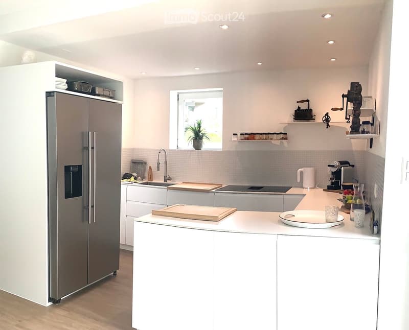 Modern kitchen with white cabinets, stainless steel refrigerator, island with wooden cutting board, coffee maker, and various kitchen items on the counter.