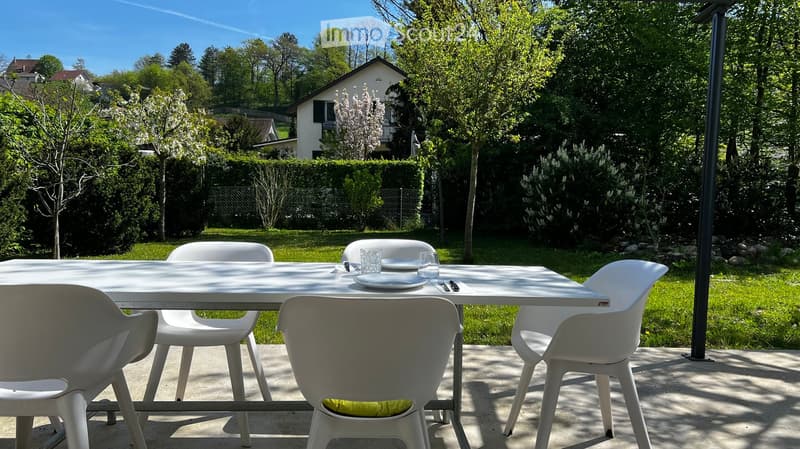 A sunny outdoor dining area with a white table and four white chairs, set on a concrete patio surrounded by a well-maintained lawn and trees.