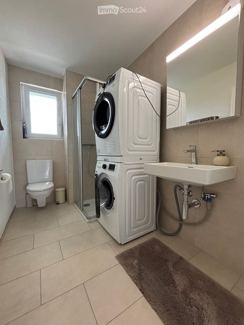 Modern bathroom with a toilet, sink, mirror, stacked washing machine and dryer, glass shower enclosure, and a small window.