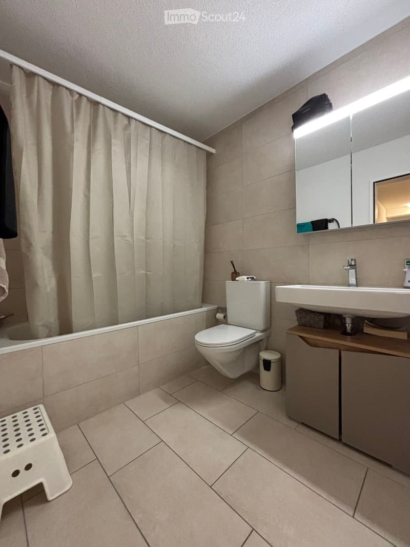 Modern bathroom with beige tiles, a bathtub, toilet, sink, mirror, and a small wooden cabinet.