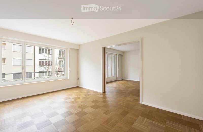 Empty room with wooden flooring, large windows, and an open doorway leading to another room.