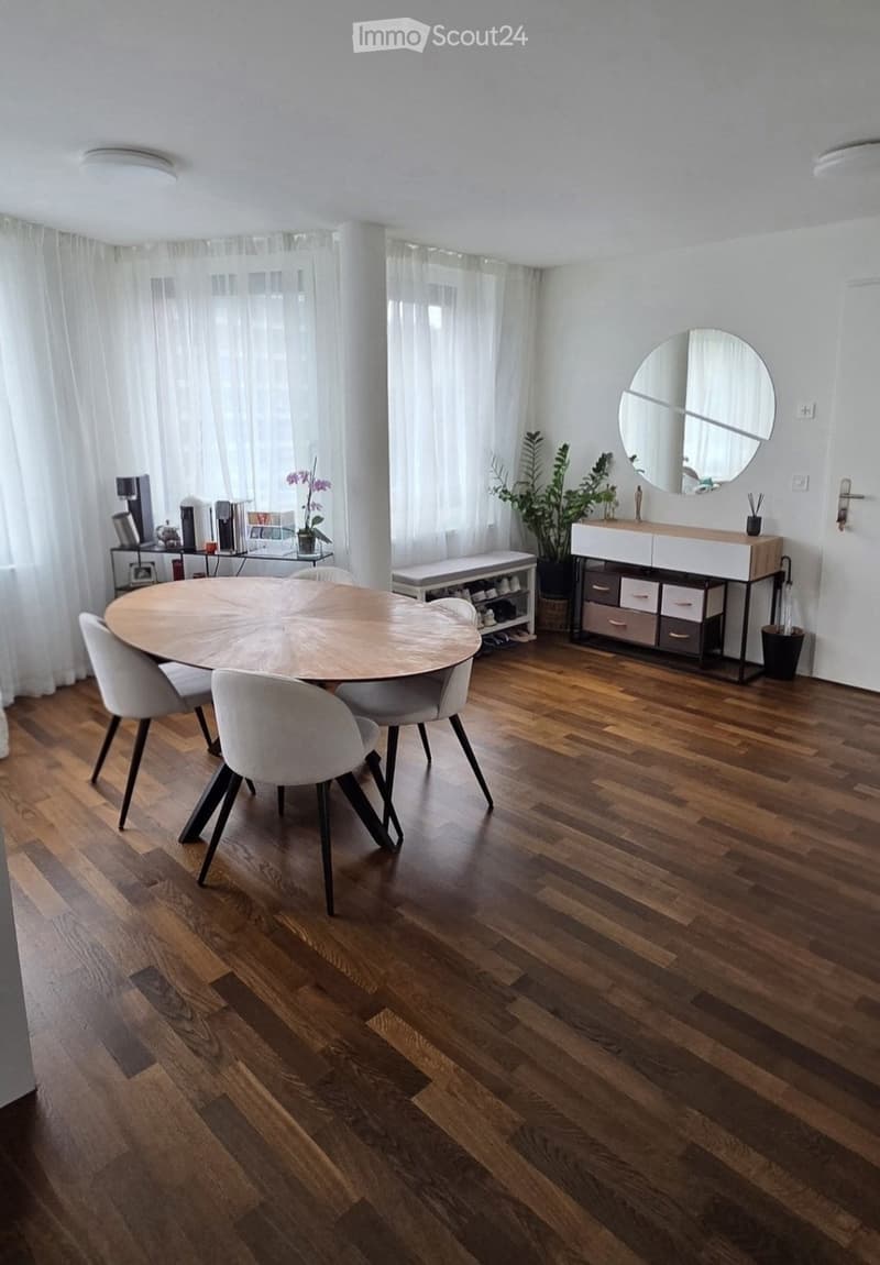 Bright dining area with wooden floor, round wooden table surrounded by white chairs, storage unit with drawers, bench, potted plants, and a round mirror on the wall.