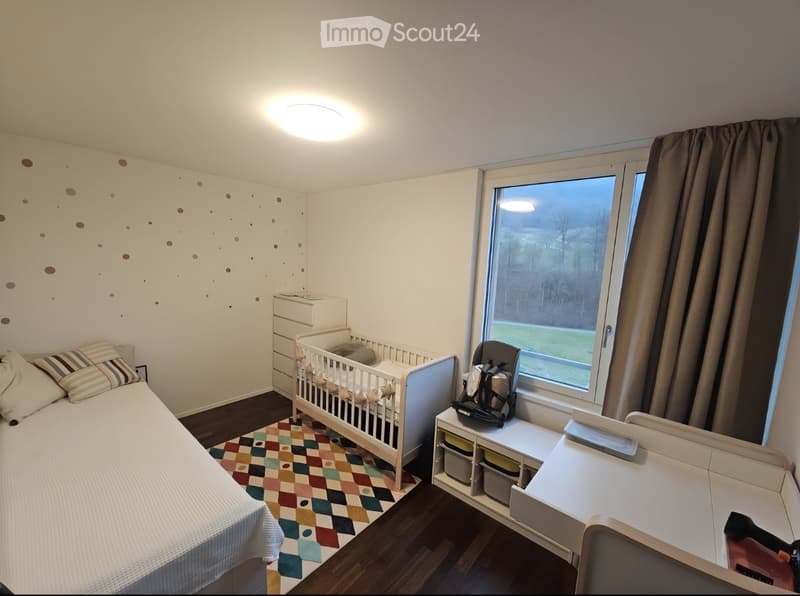 Cozy bedroom with a single bed, crib, nightstand, and a colorful rug. Features a large window with a view of the outdoors.