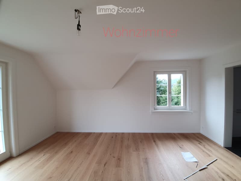 Empty living room with wooden flooring, white walls, and a single window showing greenery outside. No furniture or equipment present.