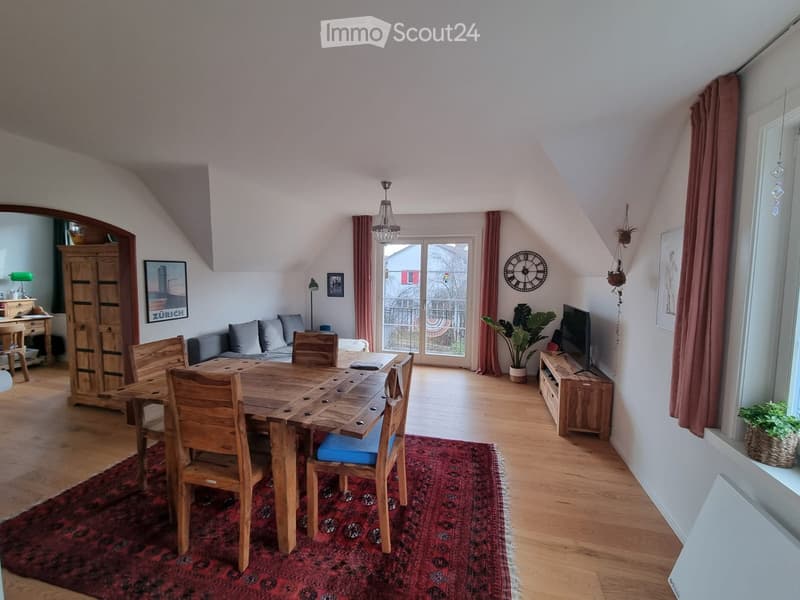 Bright, open living and dining area with wooden floors, a large dining table with chairs, a sofa, a TV stand, and decorative elements including a clock and artwork.