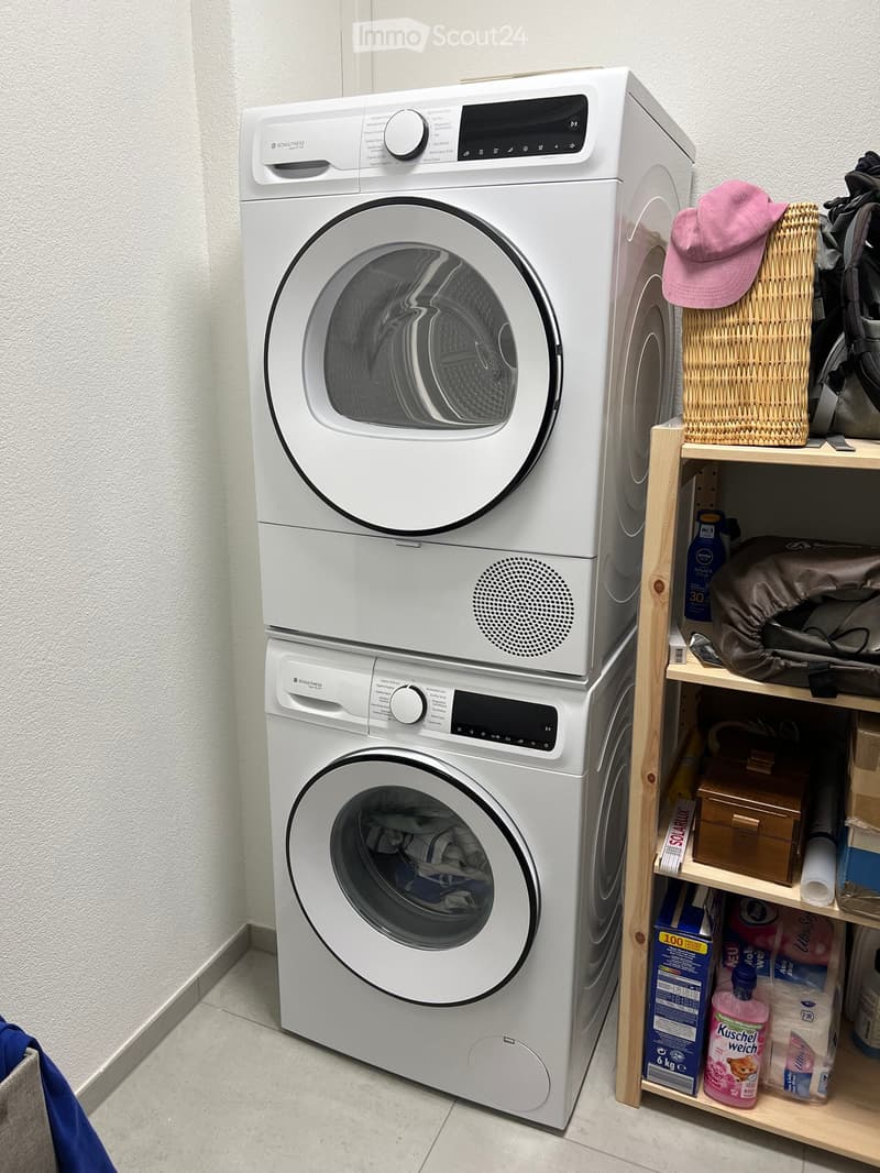 A modern bathroom with a white washing machine and dryer stacked vertically, a wooden shelf holding various items including a pink hat, a basket, and cleaning supplies.
