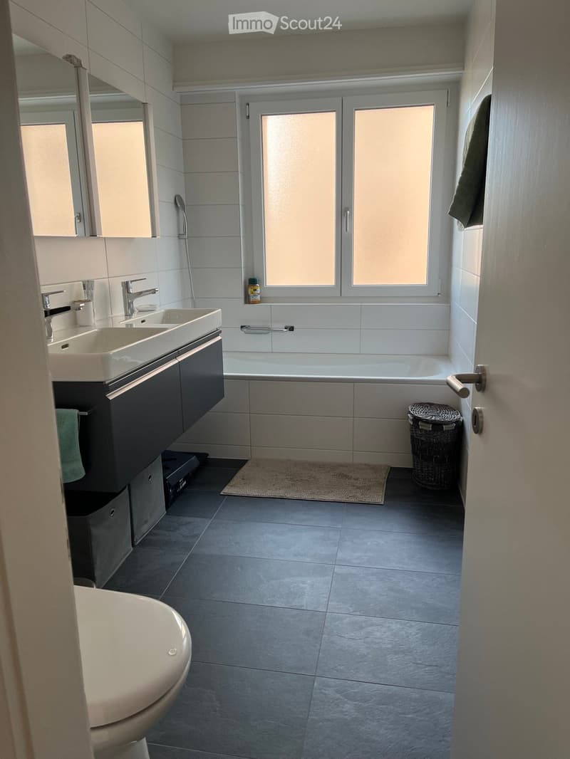 Modern bathroom with double sink, bathtub, toilet, large mirror, and wicker basket on floor.