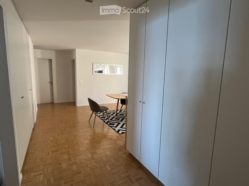 A small, modern room with wooden flooring, a single chair, and a small table. The room has white walls, a large mirror, and a patterned rug.