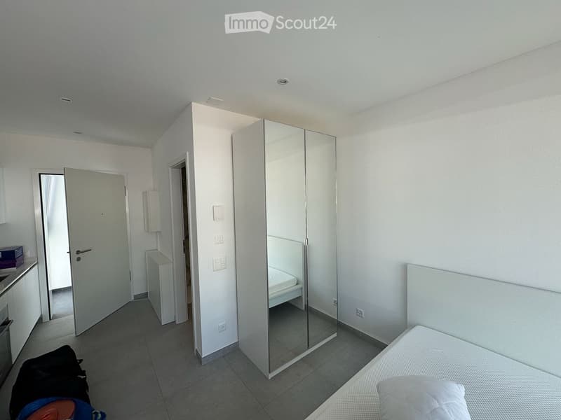 Modern, minimalist bedroom with a single bed, large mirrored wardrobe, and tiled floor. The room features white walls and a neutral color palette.