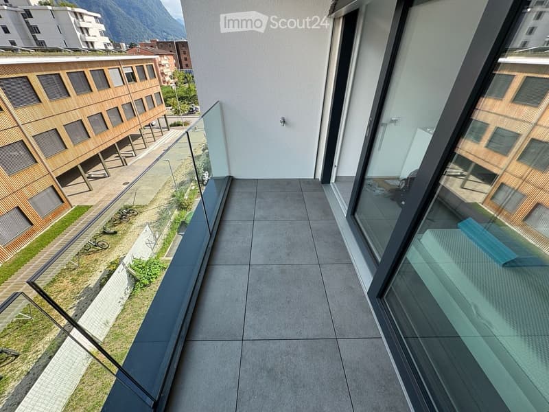 Modern balcony with large glass railings, tiled floor, overlooking a landscaped area with wooden structures and greenery.