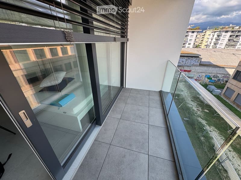 Modern balcony with large glass windows, tiled floor, and a view of the surrounding area with buildings and greenery.