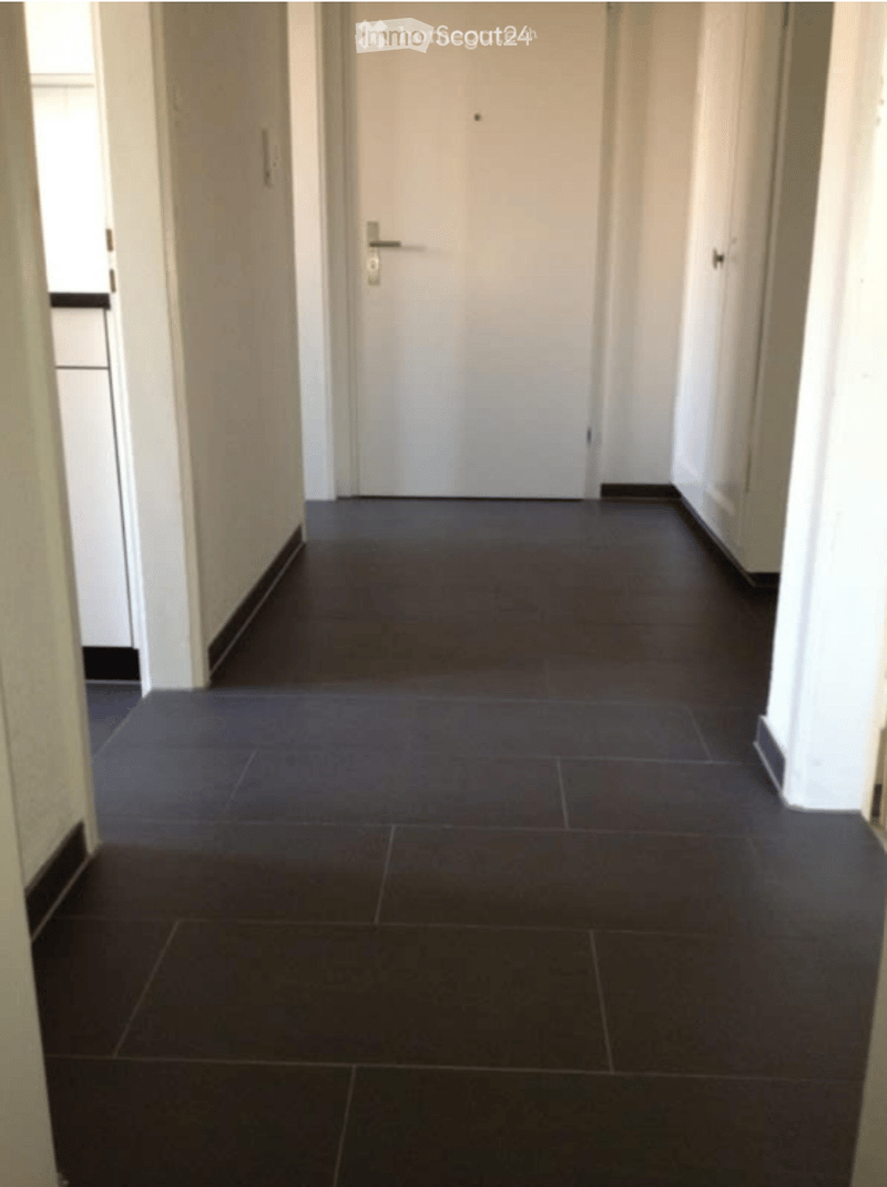 A clean, narrow hallway with dark tiled flooring and white walls. A closed white door is visible at the end of the corridor.