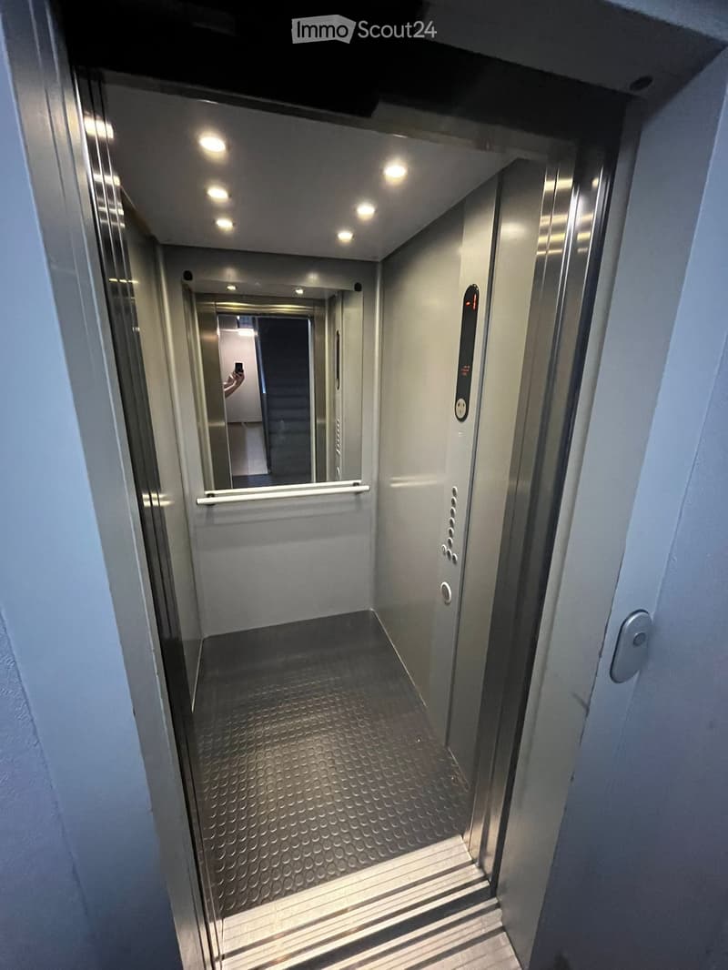 Elevator shaft with metallic walls, control panel, and illuminated ceiling lights. The elevator doors are open, revealing a clean, modern interior with a textured floor and handrail.