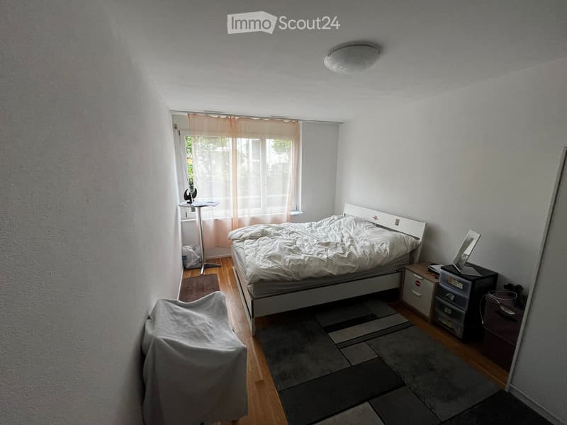 Cozy bedroom with a bed, a covered chair, a small table near the window, a mirror, and storage units.