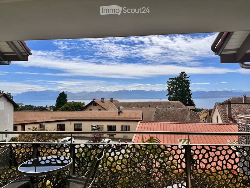 Balcony with a view of a town, lake, and mountains. Features a table and chair, with a decorative metal railing.