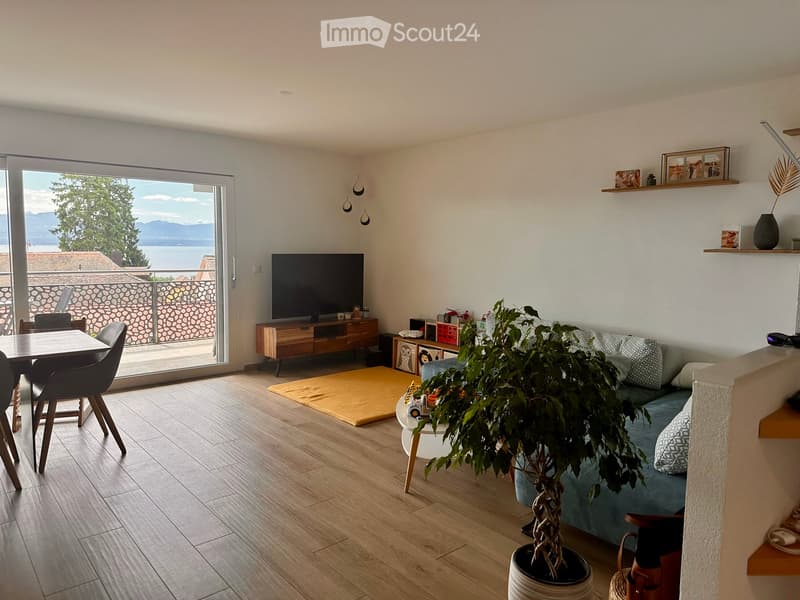 Modern living room with wooden flooring, large window offering a scenic view, sofa, dining table, chairs, television, and decorative plant.
