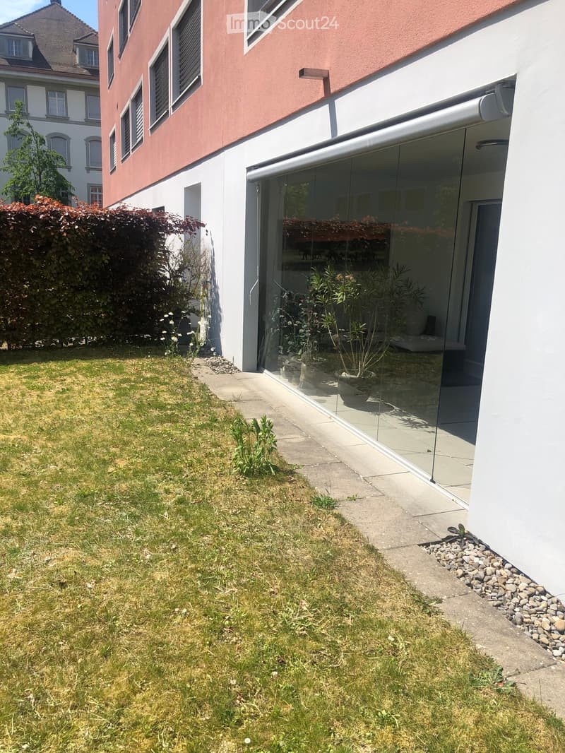 Garden area with grass, small plants, and a modern building with large glass windows and a white facade.