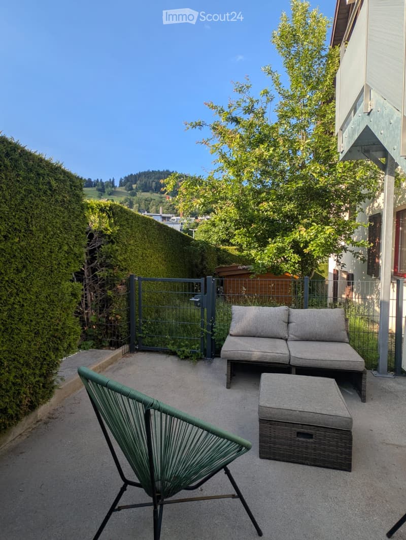 A small outdoor patio with a grey sofa, a green chair, and a small table, surrounded by greenery and a metal gate.