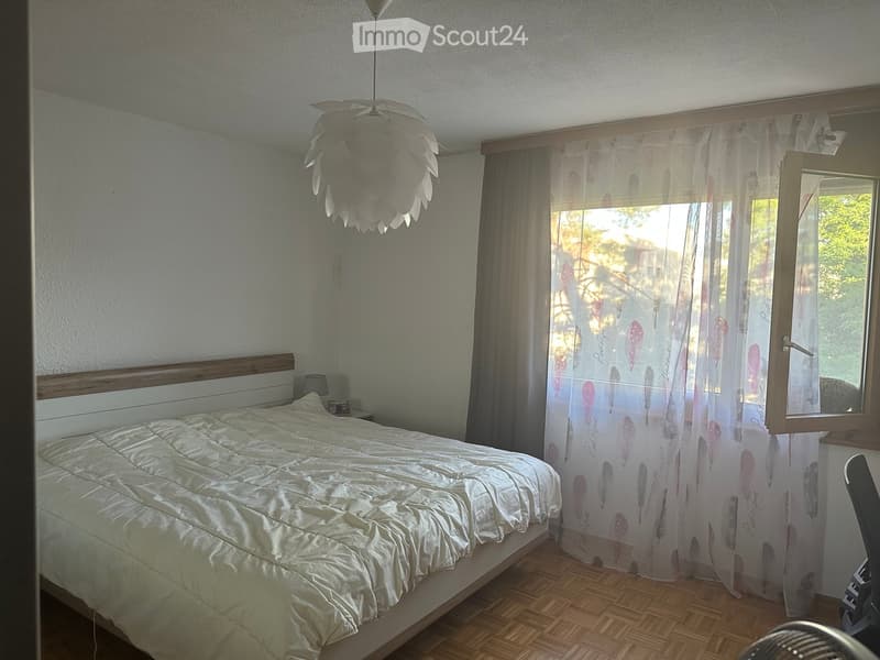 A well-lit bedroom with a large bed, wooden nightstand, and a decorative hanging light fixture. The room features wooden flooring and sheer curtains with a feather design.