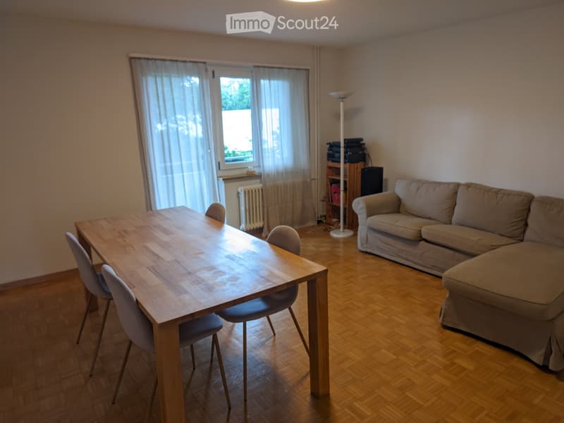 A modern living room with a wooden table, four chairs, a beige sofa, a floor lamp, and a small shelf with electronic devices.