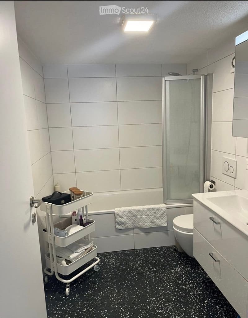 Modern bathroom with white tiles, black speckled floor, bathtub, toilet, sink, and a white cart holding towels and toiletries.