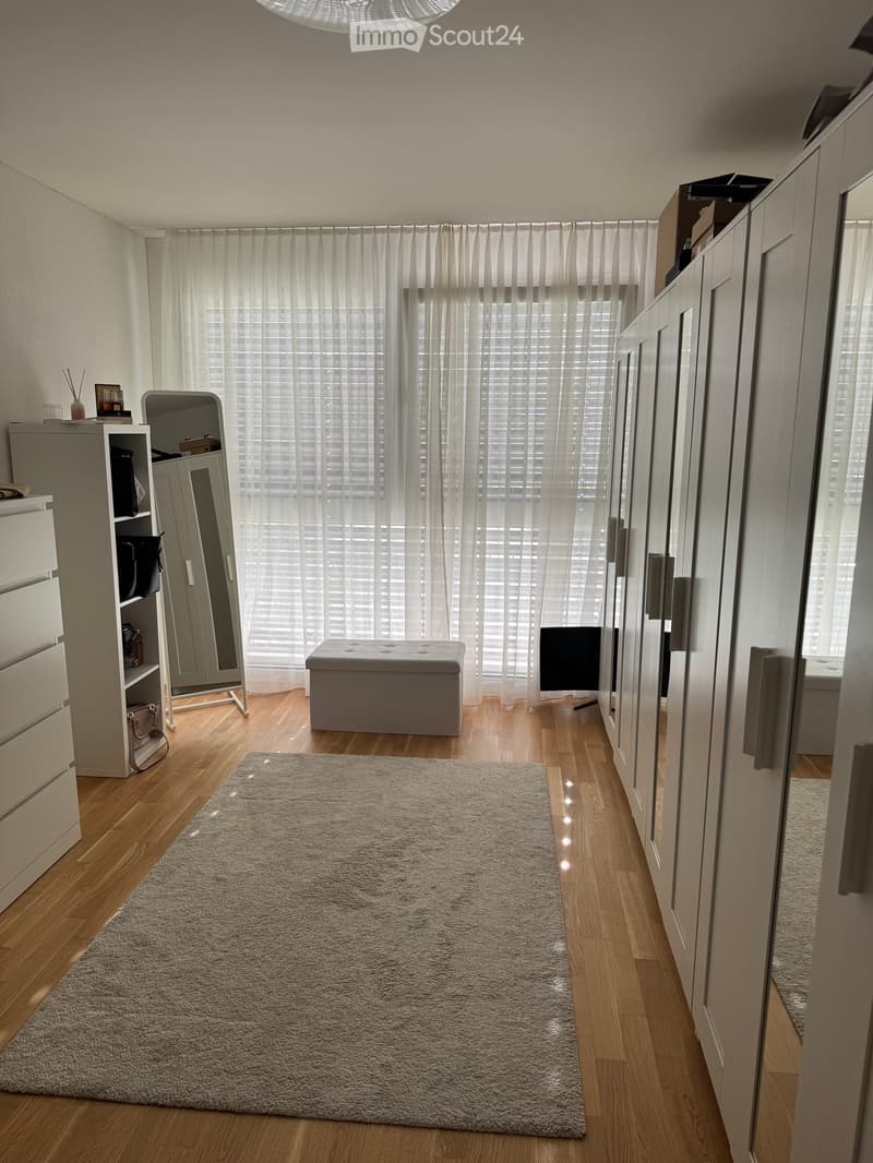 A well-lit room with wooden flooring, a large window with sheer curtains, a grey rug, a white stool, a full-length mirror, and a wardrobe with shelves.
