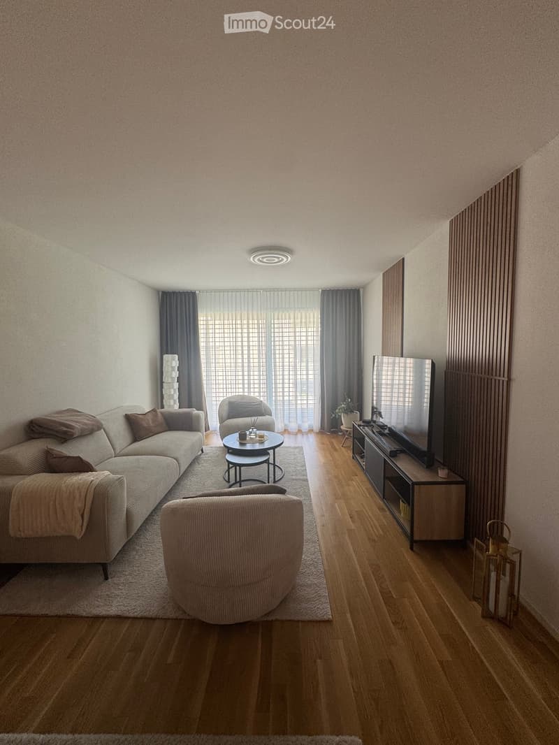Modern living room with a light-colored sofa, armchair, coffee table, TV stand, and a round table with chairs. The room features wooden flooring, large windows with curtains, and a cozy rug.