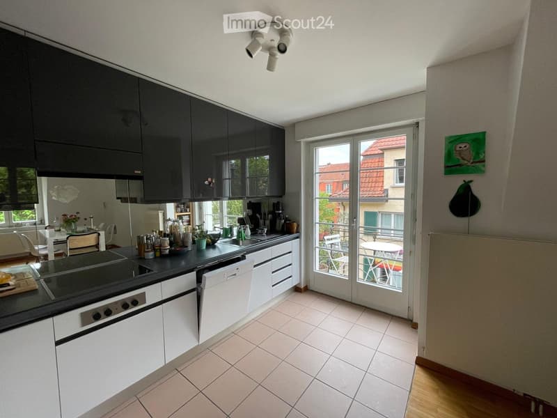 Modern kitchen with white cabinets, black countertops, and stainless steel appliances. Features an oven, dishwasher, sink, and various kitchen items on the counter. A sliding glass door leads to a balcony with outdoor furniture.