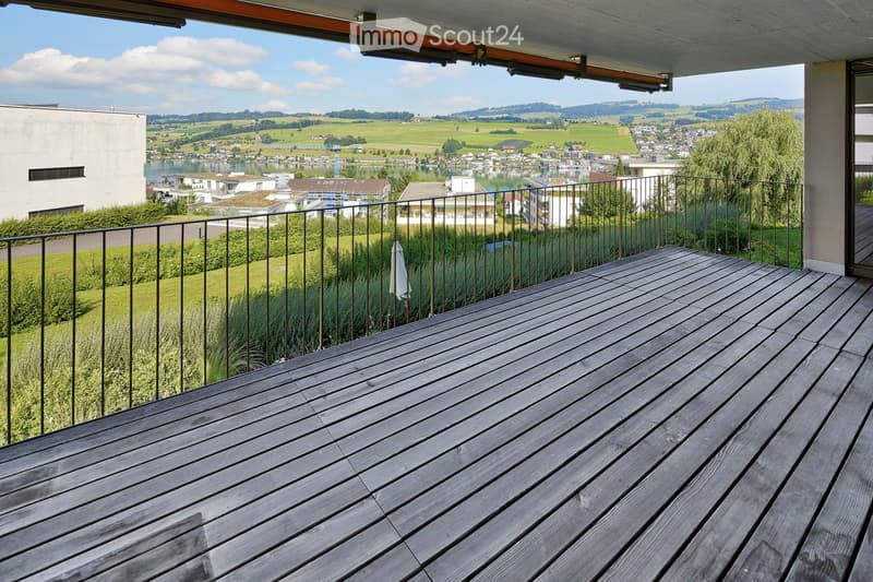 Wooden deck with metal railing, overlooking a scenic landscape with fields, water, and distant hills.