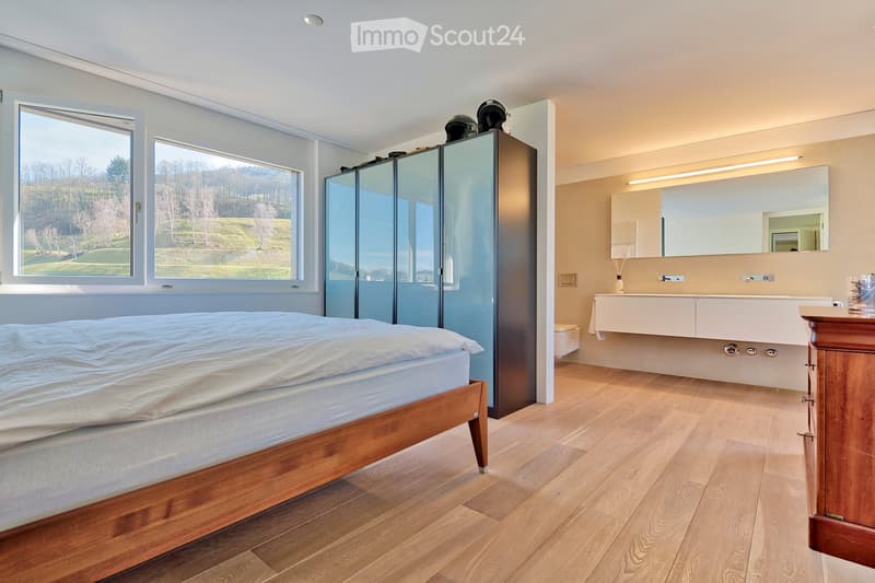 Spacious bedroom with large windows offering a scenic view, featuring a wooden bed, modern wardrobe, and an en-suite bathroom with a sink and mirror.