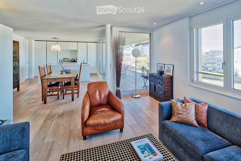 Modern living and dining area with hardwood floors, large windows offering a view, a blue sofa with pillows, a leather armchair, a coffee table, a dining table with chairs, a floor lamp, a decorative cabinet, and a tall vase with dried plants.