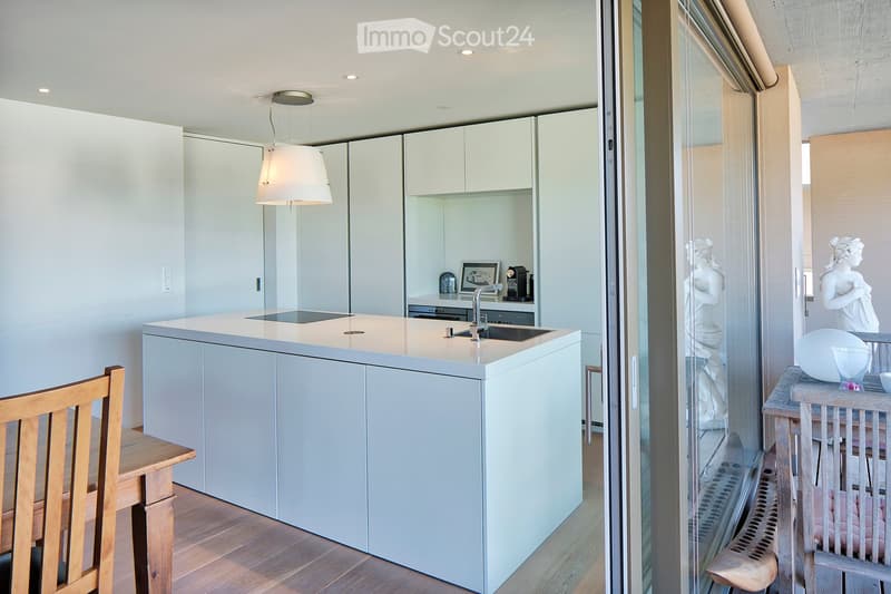 Modern kitchen with white cabinets, a central island, and a sink. A wooden chair and table are visible, along with a hanging lamp and a view of a balcony area with decorative items.