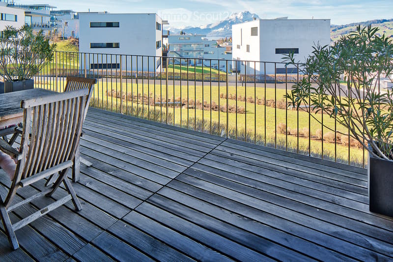 Wooden balcony with a single wooden chair, metal railing, and potted plants, overlooking a green field and distant mountains.
