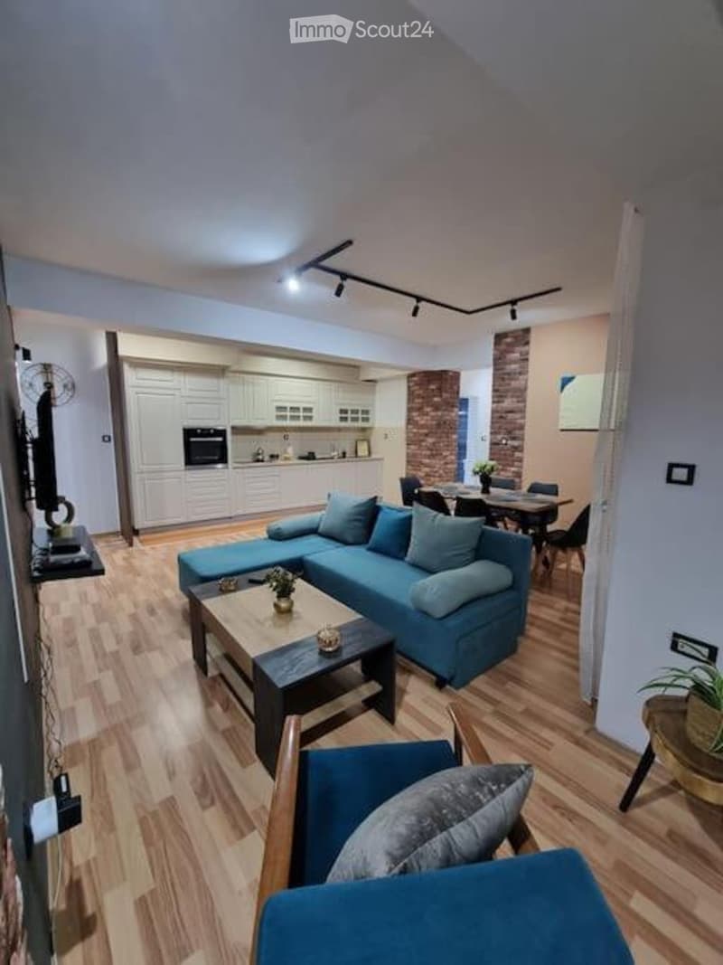 Modern living room with a blue sectional sofa, coffee table, dining area with chairs and table, kitchen with white cabinets and an oven, and a fan near the TV.