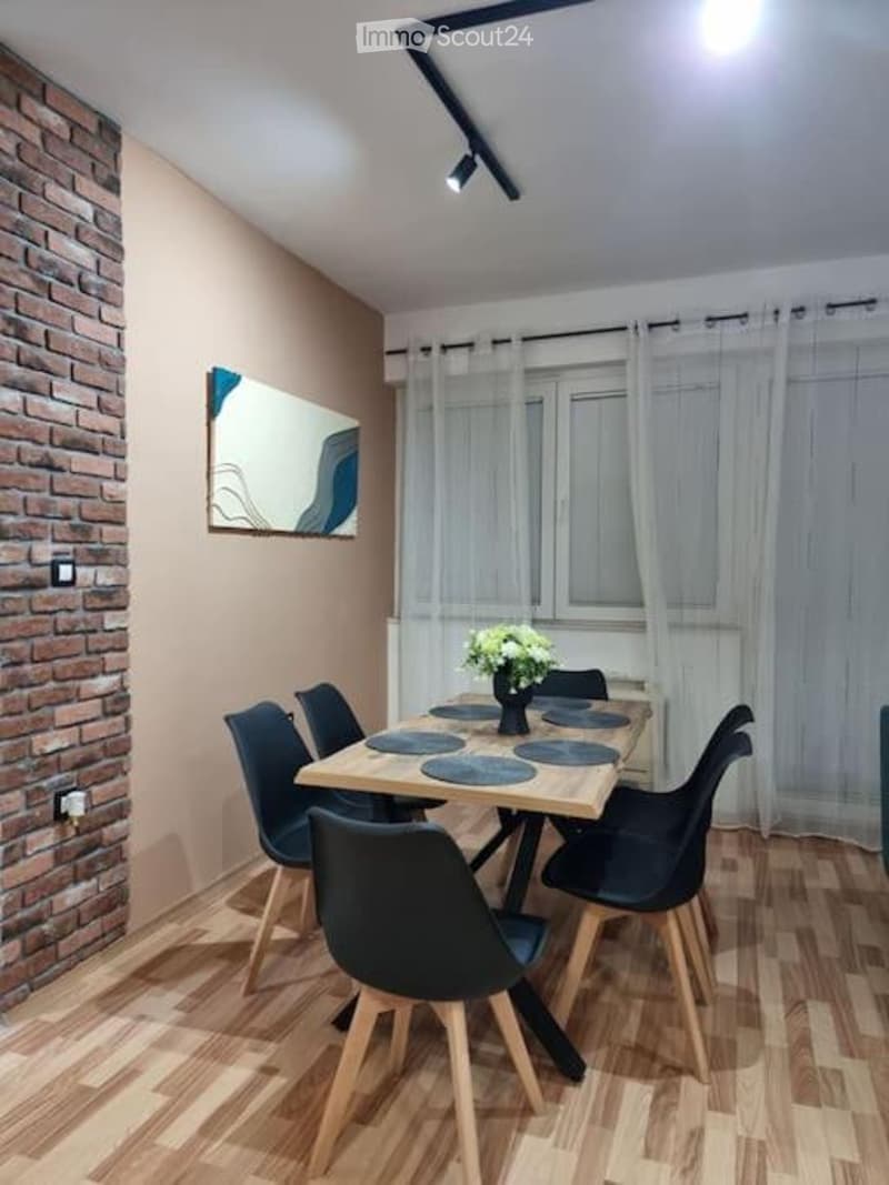 Dining room with wooden floor, brick accent wall, large window with white curtains, dining table with six chairs, flower vase, and artwork on wall.