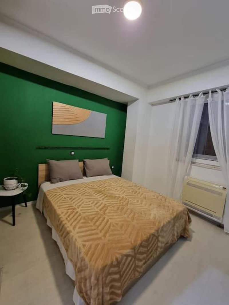 Cozy bedroom with a double bed, nightstand, small table, and artwork on the wall. The room features green and white walls, a large window with white curtains, and a warm, inviting atmosphere.