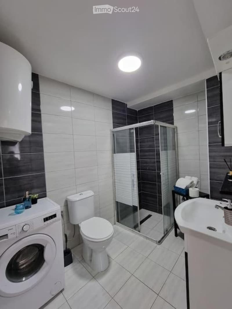 Modern bathroom with white tiles, a toilet, sink, shower, washing machine, and a small shelf with towels.
